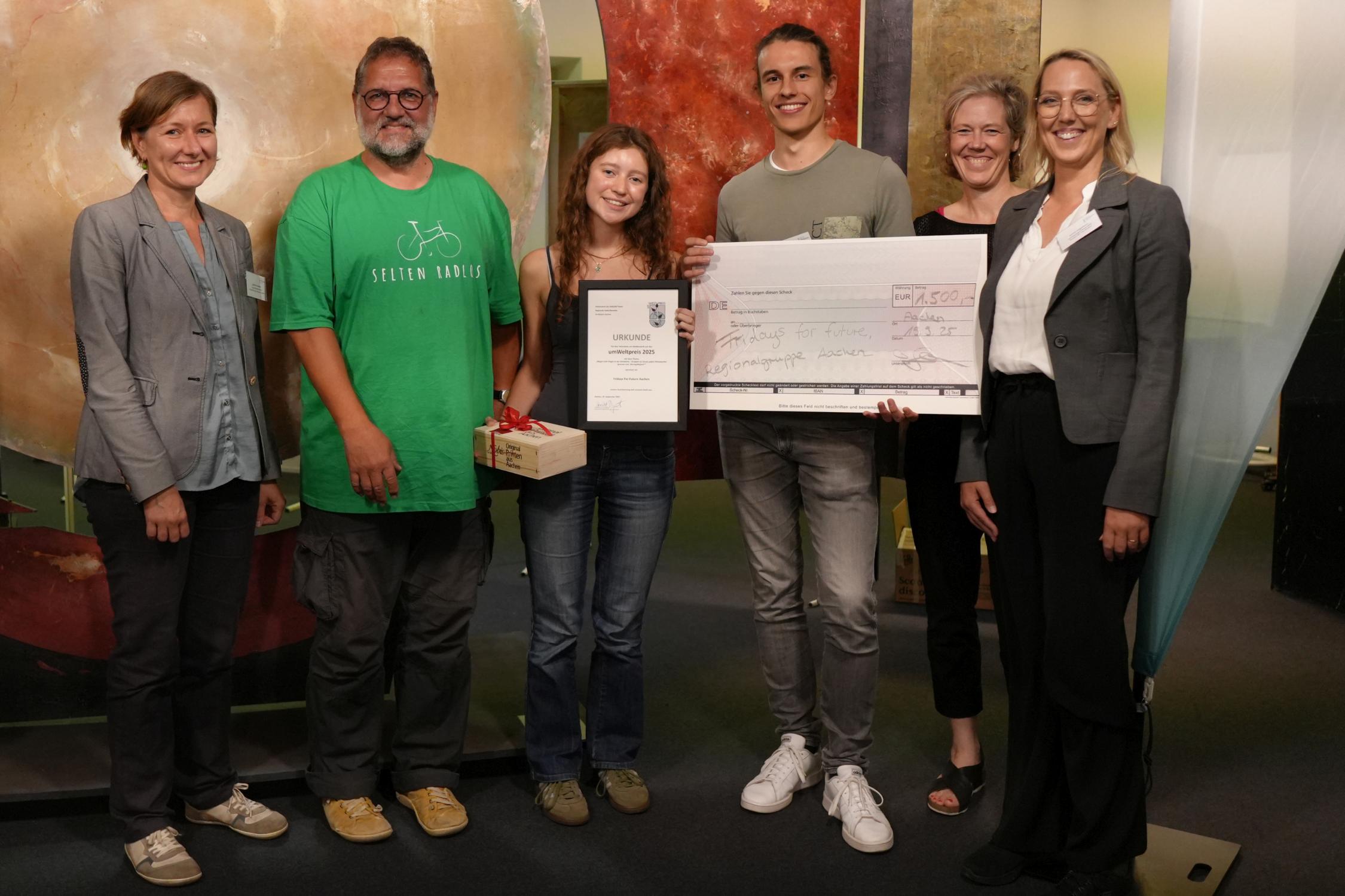 Vier Frauen und zwei Männer. Eine Frau hält eine Urkunde in der Hand und eine Schachtel. Ein Mann hält einen großen Scheck über 1.500 Euro in der Hand.